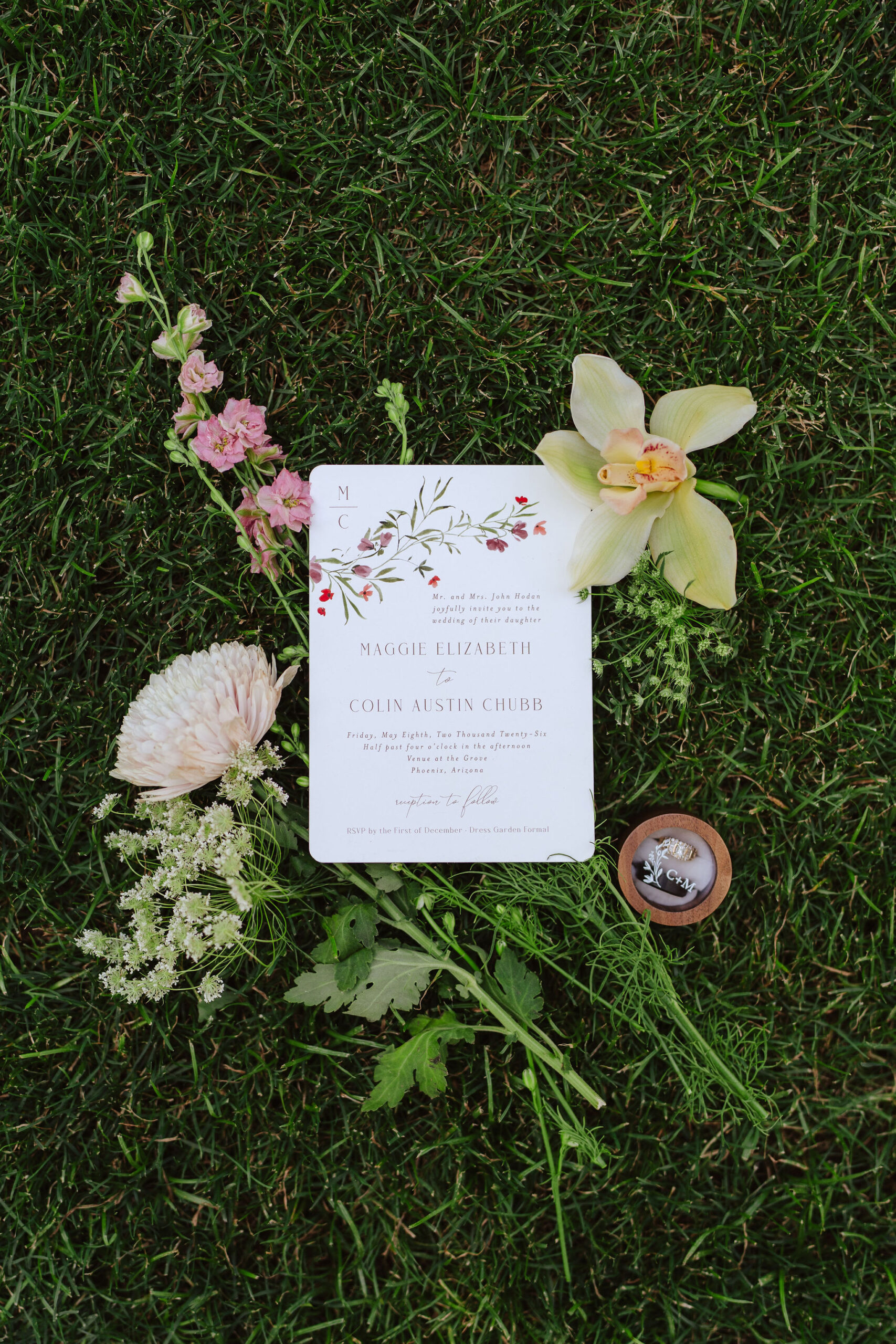 **Alt text:** A wedding invitation for Maggie Elizabeth and Colin Austin Chubb, dated Friday, May 8th, 2026, at Venue at the Grove in Phoenix, Arizona, displayed on green grass surrounded by pink stock flowers, a cream chrysanthemum, a green cymbidium orchid, Queen Anne's lace, and a small wooden ring box with wedding rings.