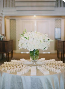 **Alt text:** *A round table with a light blue tablecloth displays rows of small white place card holders arranged in concentric arcs, centered around a glass vase filled with white hydrangeas and lilies. The elegant setting suggests a wedding reception in a softly lit, neutral-toned venue.*