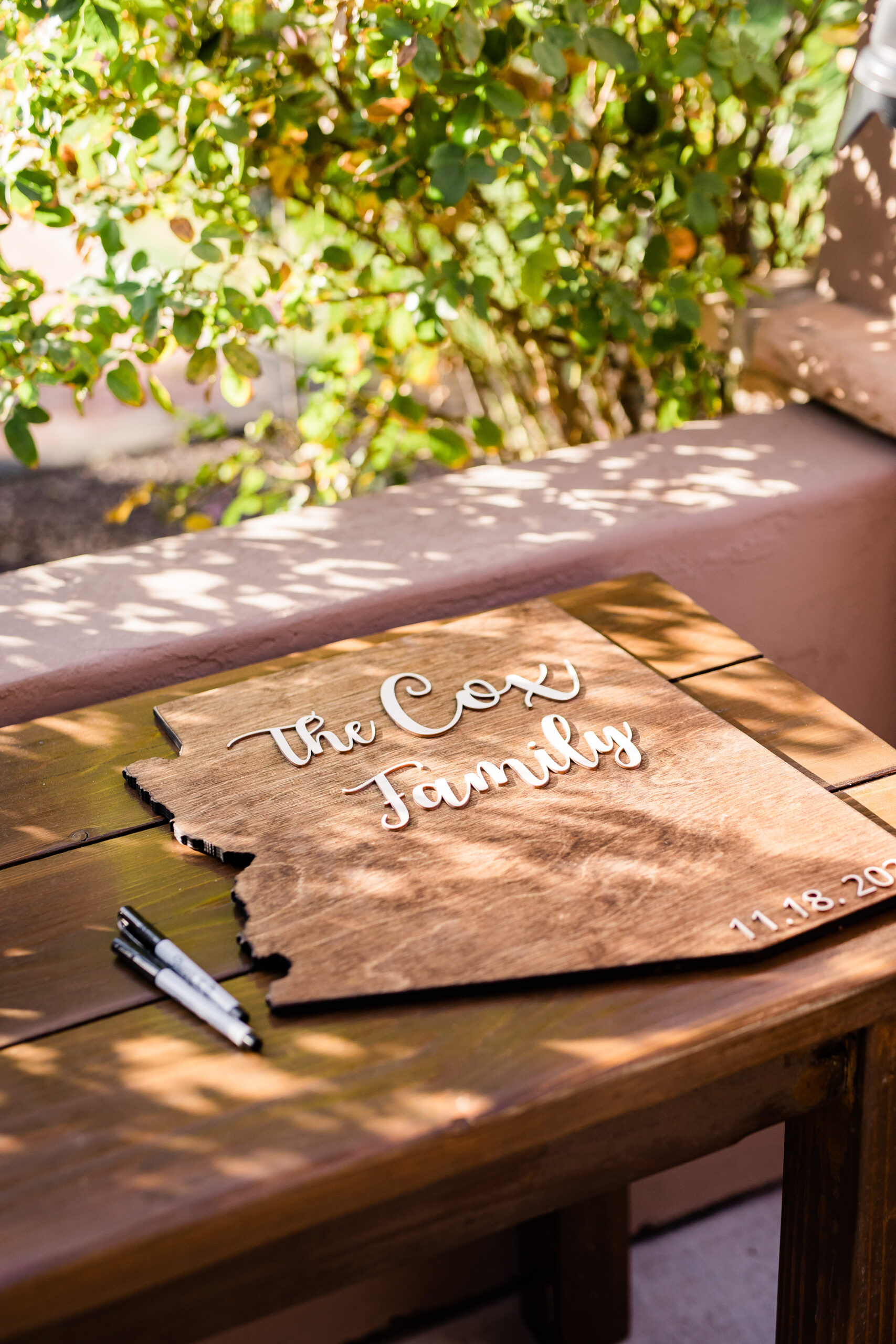 **Alt text:** A wooden guest book shaped like the state of Arizona rests on a picnic table outdoors, engraved with white script reading "The Cox Family" and the date "11.18.20" in the corner. Two black markers lie beside it, with dappled sunlight and green foliage visible in the background.
