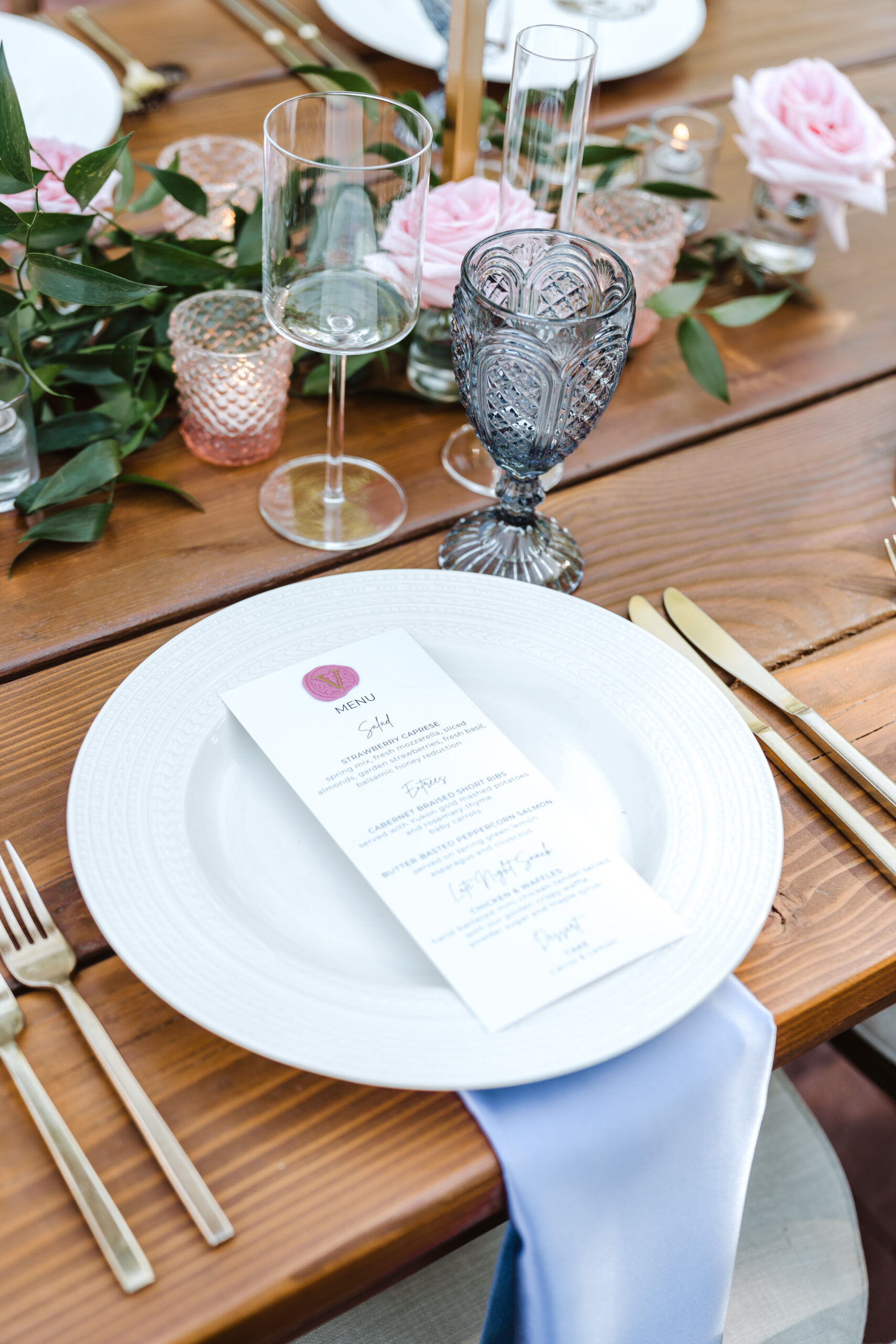 **Alt text:** An elegant wedding reception place setting on a rustic wood table, featuring a white ribbed charger plate with a printed menu card sealed with a pink wax seal, a periwinkle blue linen napkin, gold flatware, a smoke-blue cut crystal goblet, and a clear wine glass. The centerpiece includes pink garden roses, eucalyptus greenery, and pink hobnail votive candle holders.