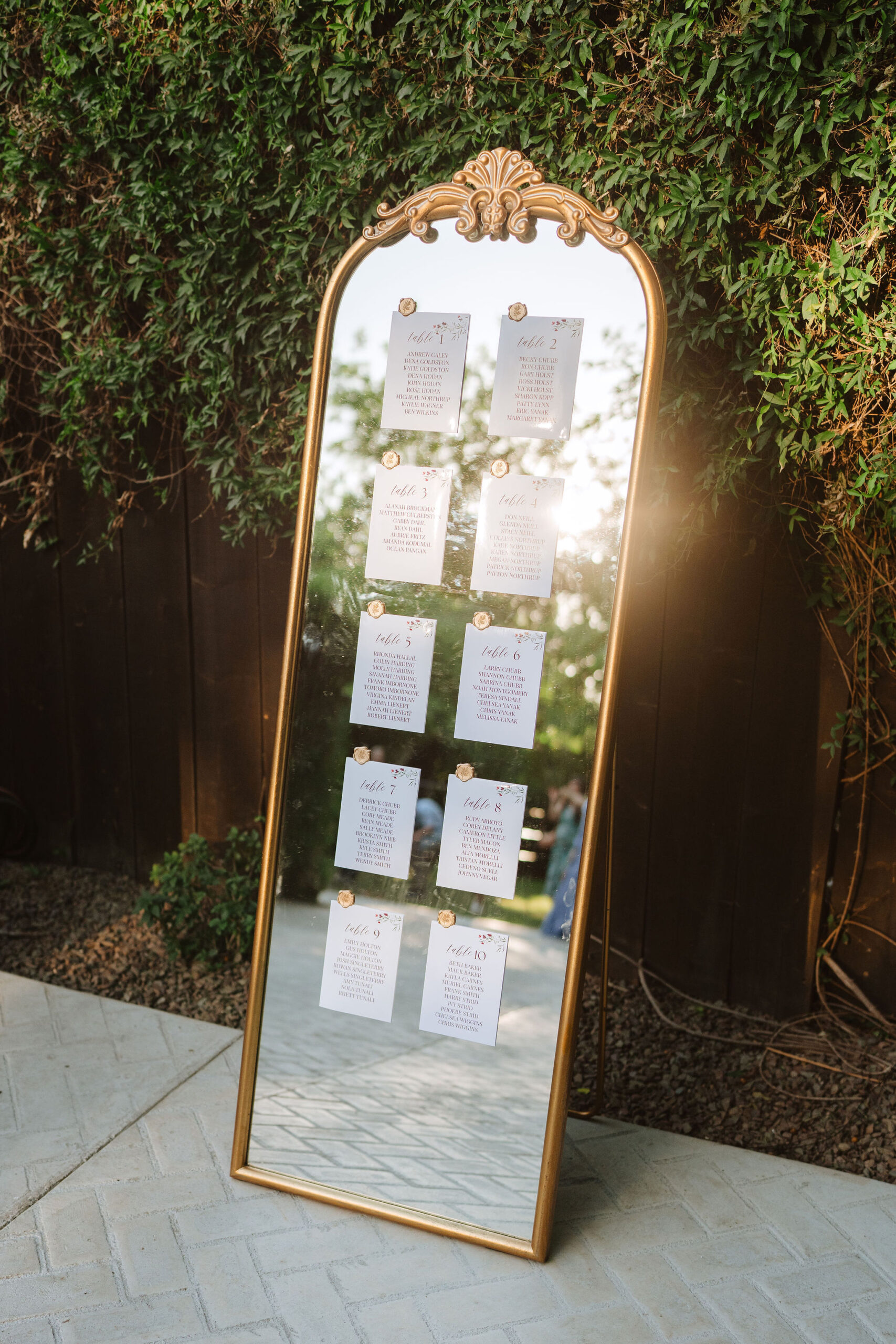 **Alt text:** A tall ornate gold-framed floor mirror leaning against a ivy-covered wall outdoors, used as a wedding seating chart display. Ten small white cards labeled "Table 1" through "Table 10," each listing guest names in calligraphy script, are attached to the mirror with gold floral clips. Warm golden sunlight reflects in the mirror.