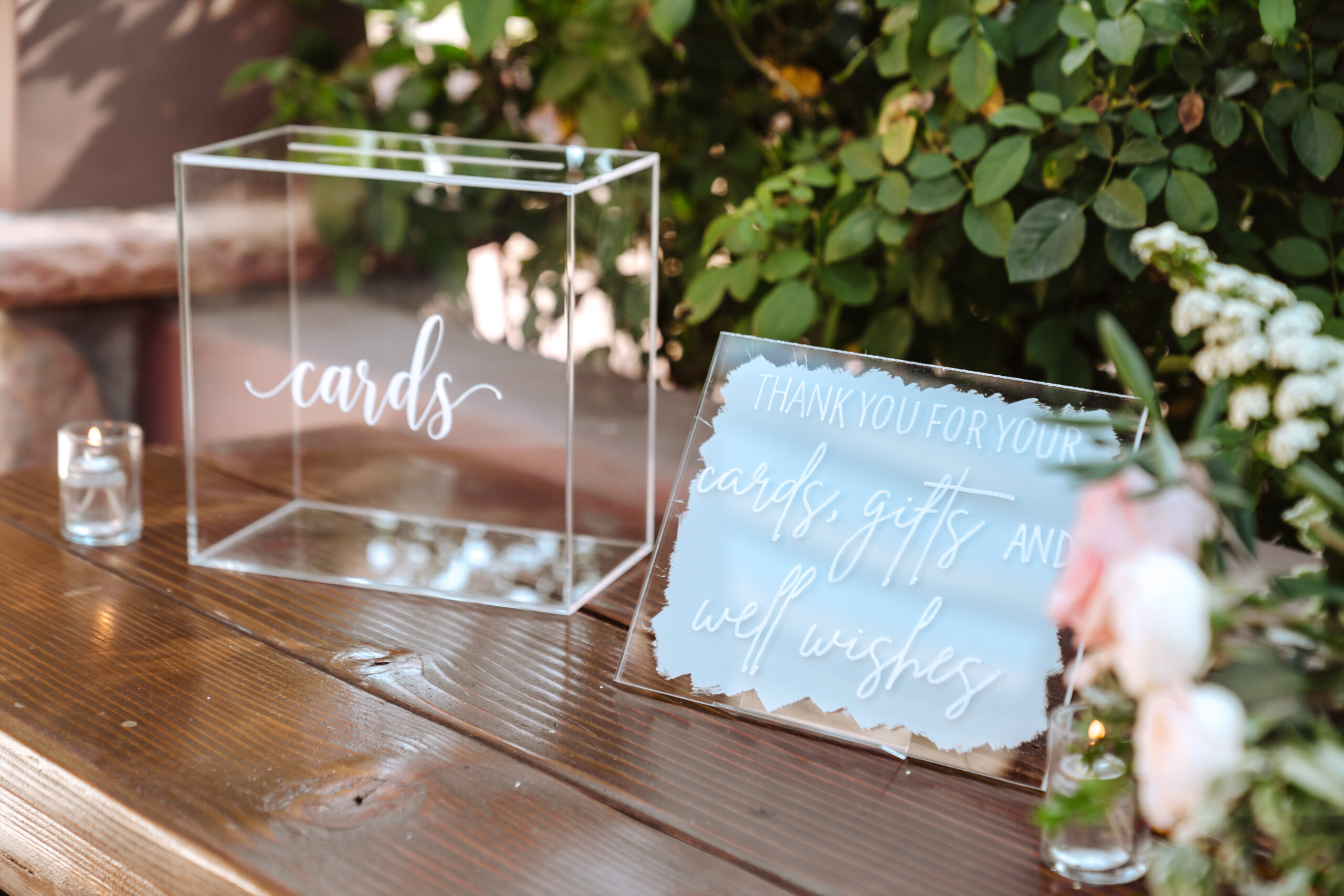 **Alt text:** A clear acrylic wedding card box labeled "cards" in white script sits on a rustic wooden table beside a mirrored acrylic sign reading "Thank you for your cards, gifts and well wishes" on a blue watercolor background. Small votive candles and pink floral arrangements with greenery accent the display.
